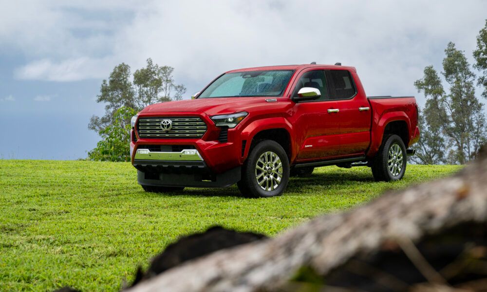 Ein Toyota Tacoma der aktuellen Generation fährt auf einer Schotterstraße, im Hintergrund eine Fabriklandschaft im Industriestil