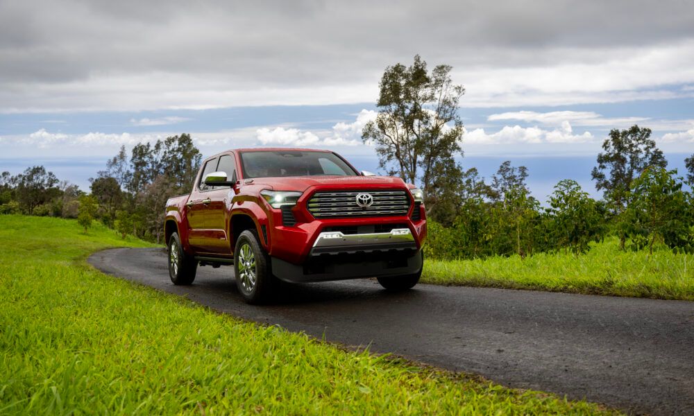 Ein Toyota Tacoma der aktuellen Generation fährt auf einer Schotterstraße, im Hintergrund eine Fabriklandschaft im Industriestil