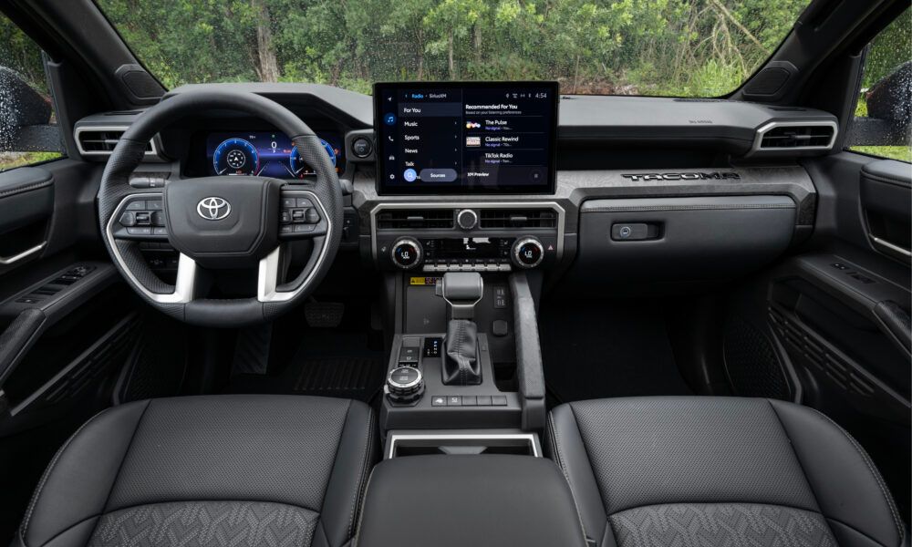 Ein Toyota Tacoma der aktuellen Generation fährt auf einer Schotterstraße, im Hintergrund eine Fabriklandschaft im Industriestil